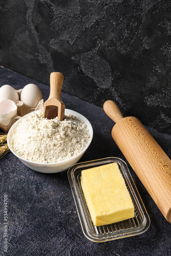 Bowl with flour, butter and rolling pin on dark table