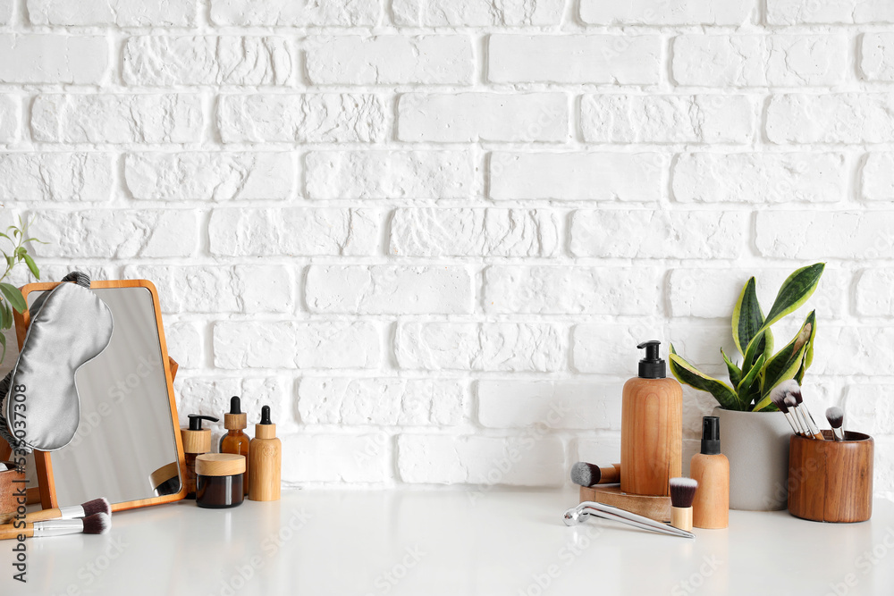 Mirror with sleeping mask, cosmetics, makeup brushes and houseplant on table near white brick wall