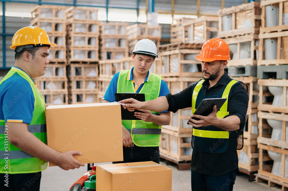 Warehouse manager wearing helmet pointing towards shelf using digital tablet and notepad