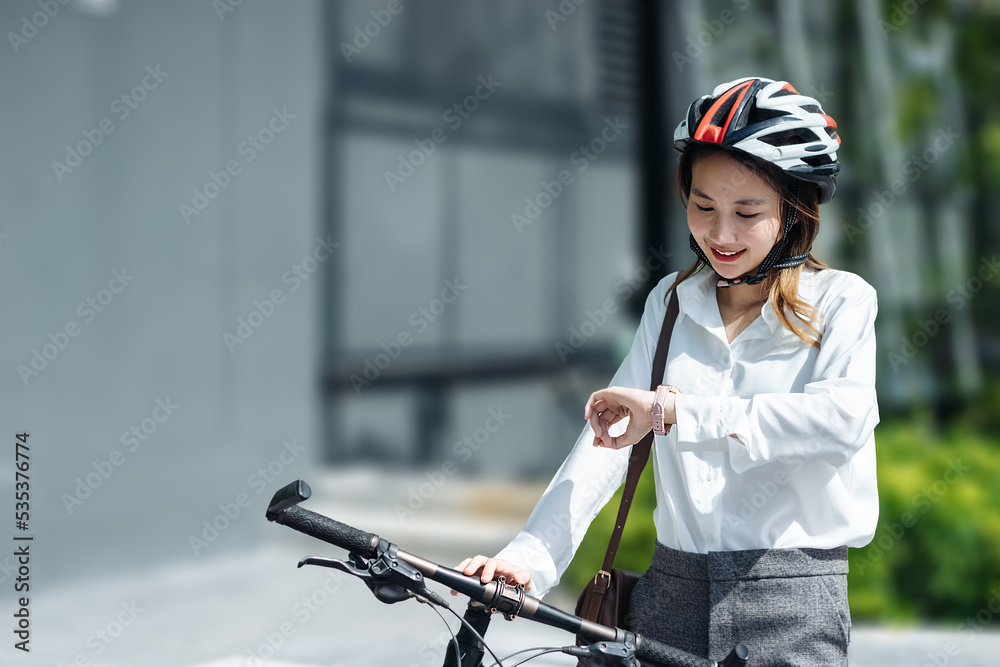 年轻的亚洲女商人戴着头盔，看着时钟，骑着自行车站在城市街道上