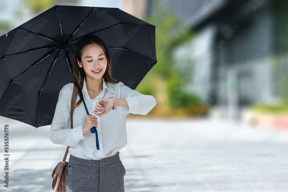 阳光明媚的日子里，打着黑色雨伞在户外看手表的可爱亚洲商业女性，专注