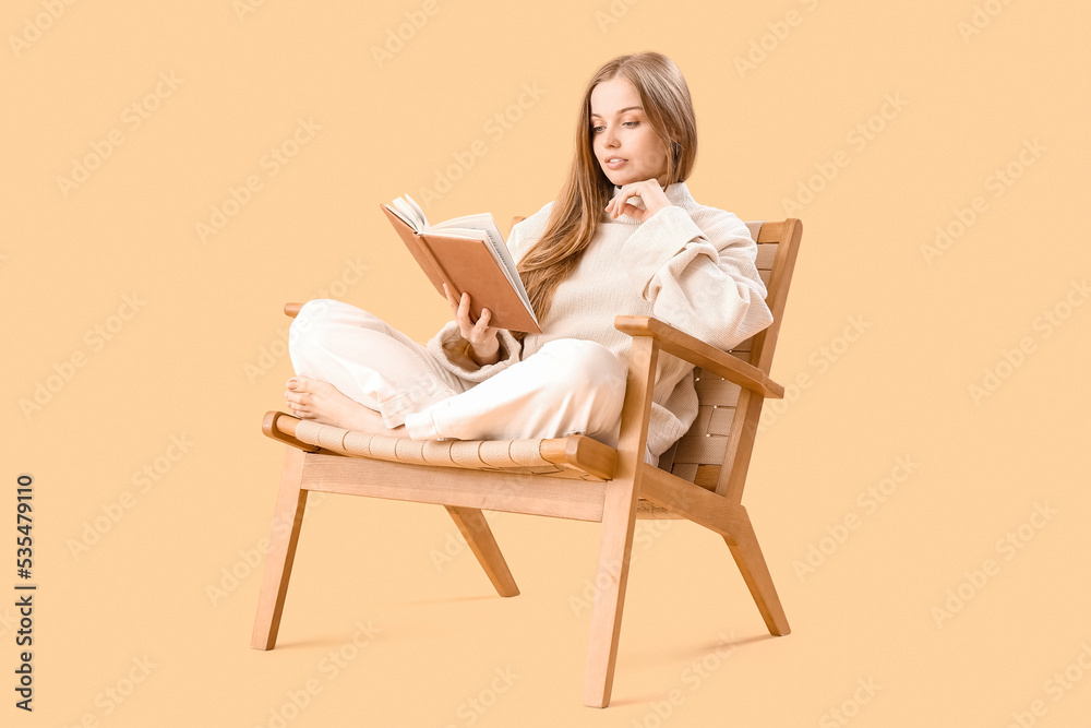 Young woman reading book in armchair on beige background