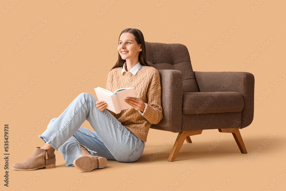 Beautiful woman reading book near armchair on beige background