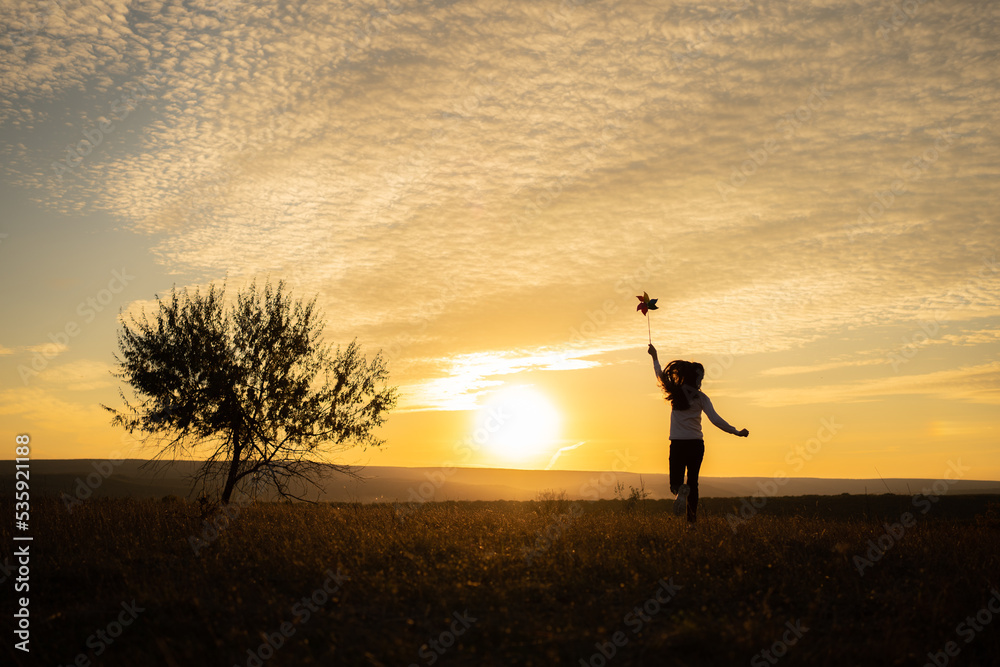 Little girl running on the meadow at sunset with windmill in her hands. Silhouette of child girl hol