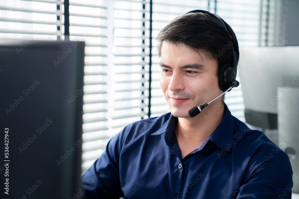 Young competent asian male call center agent working at his computer while simultaneously speaking w