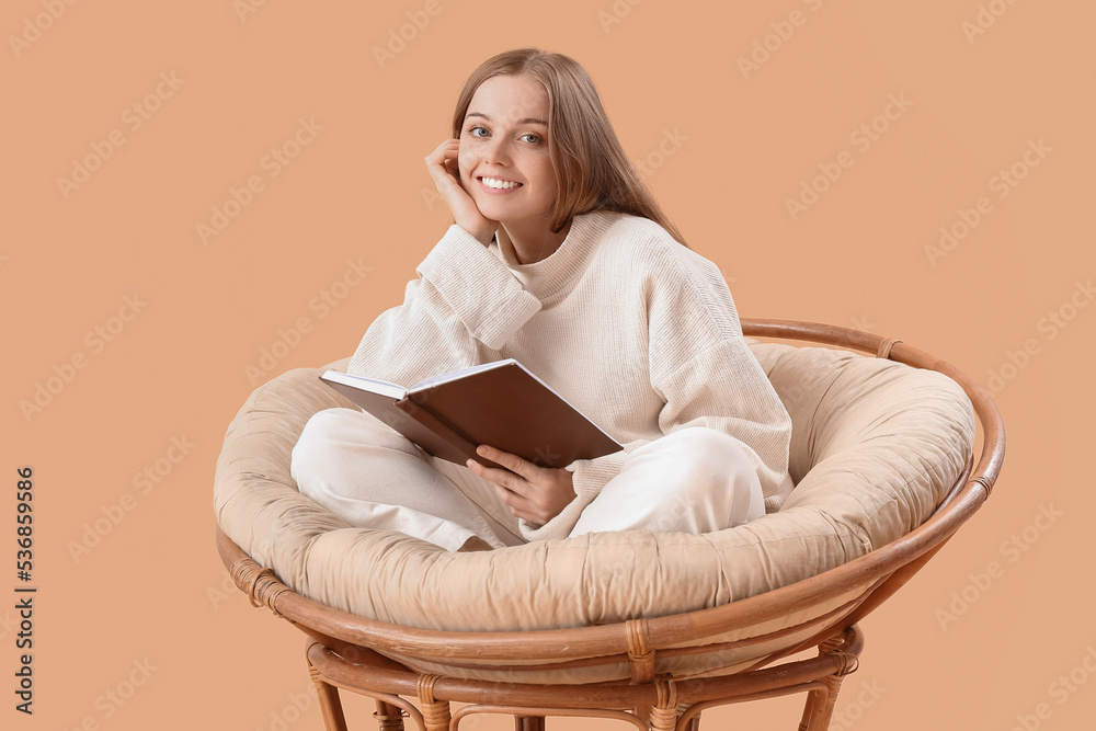 Young woman with book sitting in armchair on beige background