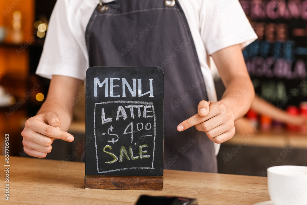 Young male barista pointing at chalkboard with menu in cafe, closeup