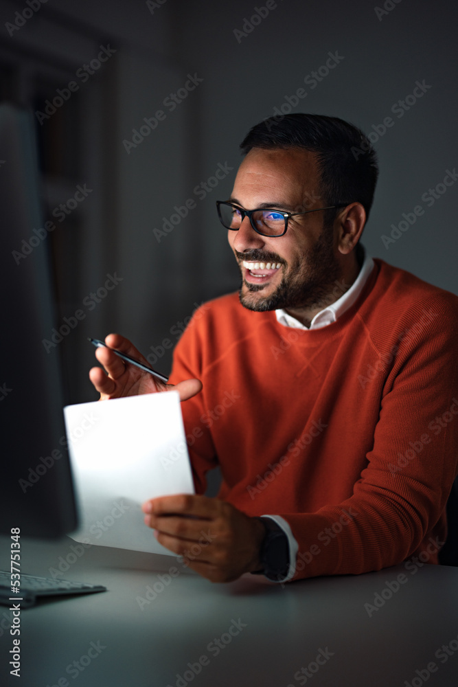 A smiling man working at the office, late at night, having an online meeting.