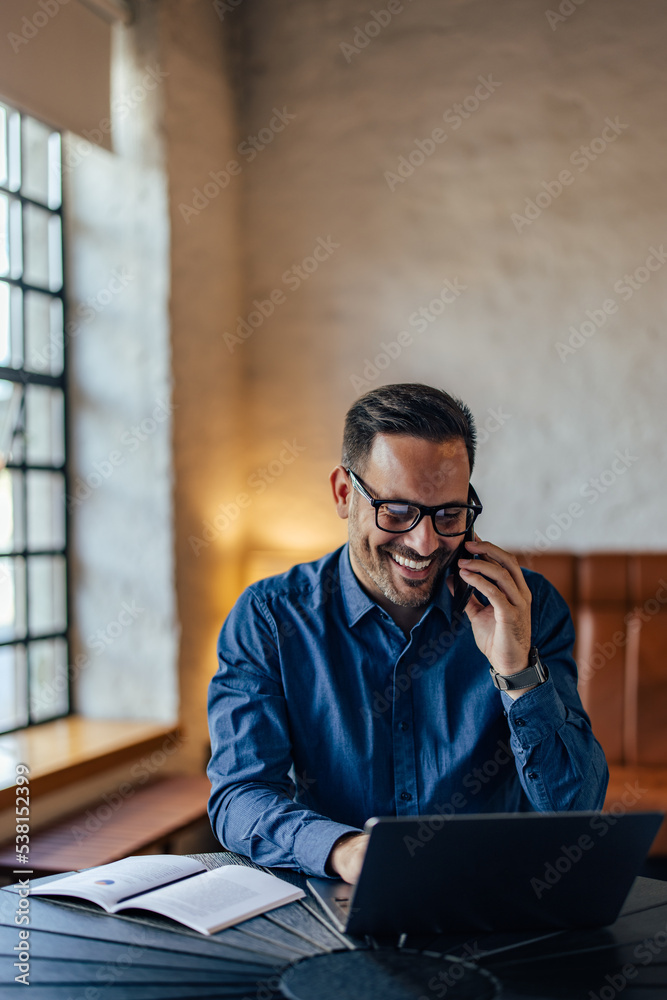 Smiling adult man with his glasses on, working over the laptop, making a phone call.