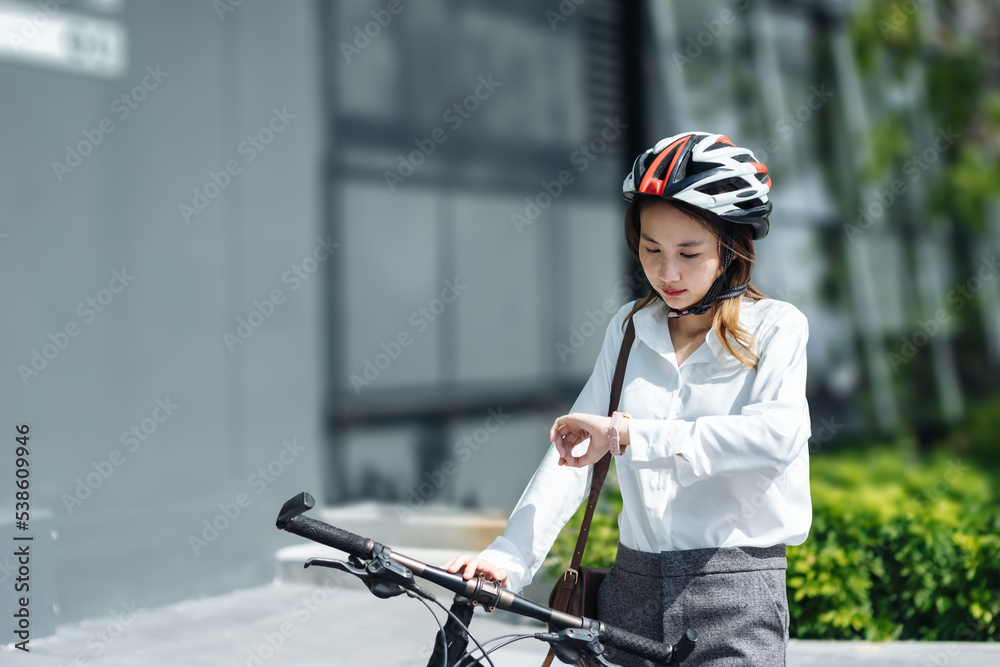 年轻的亚洲女商人戴着头盔，看着时钟，骑着自行车站在城市街道上