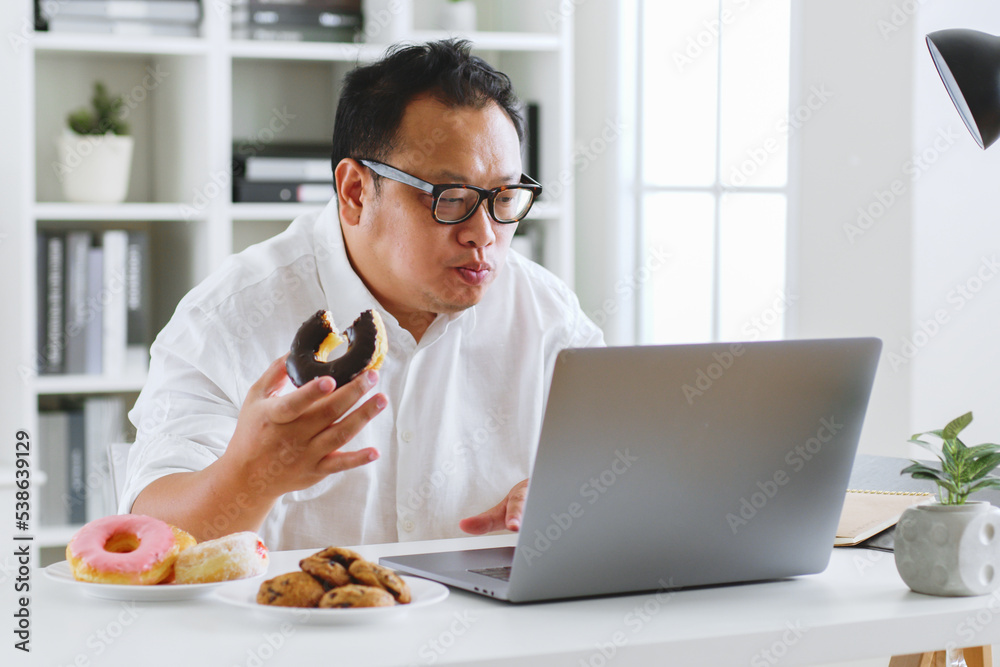 亚洲胖子在使用笔记本电脑工作时吃甜甜圈、甜食、垃圾食品、不健康饮食c