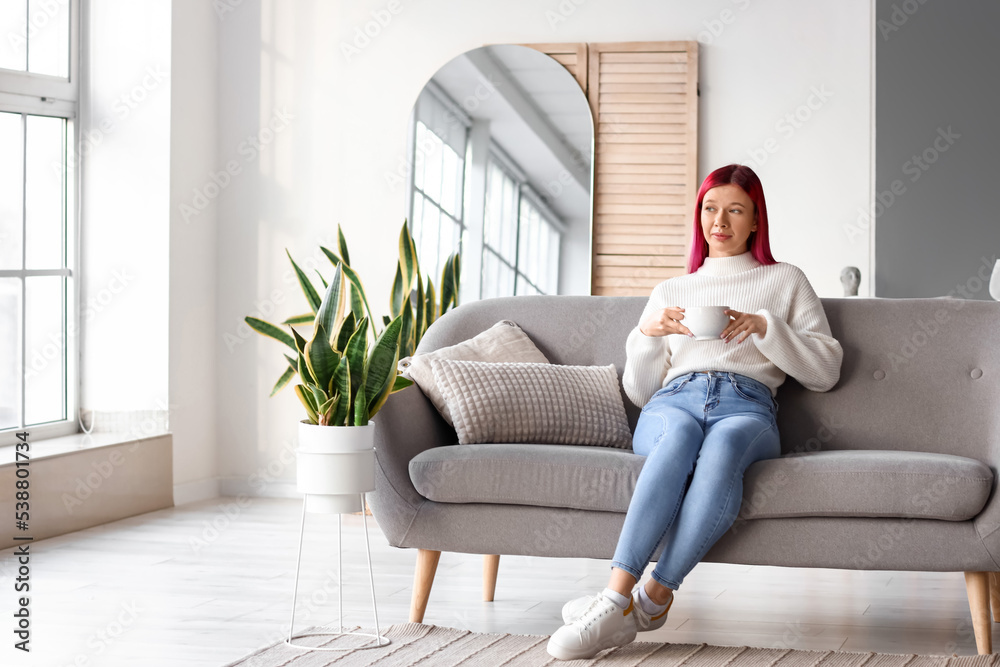 Pretty young woman with unusual hair drinking coffee while relaxing on sofa at home