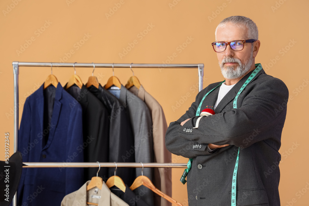 Mature fashion designer and rack with clothes on beige background