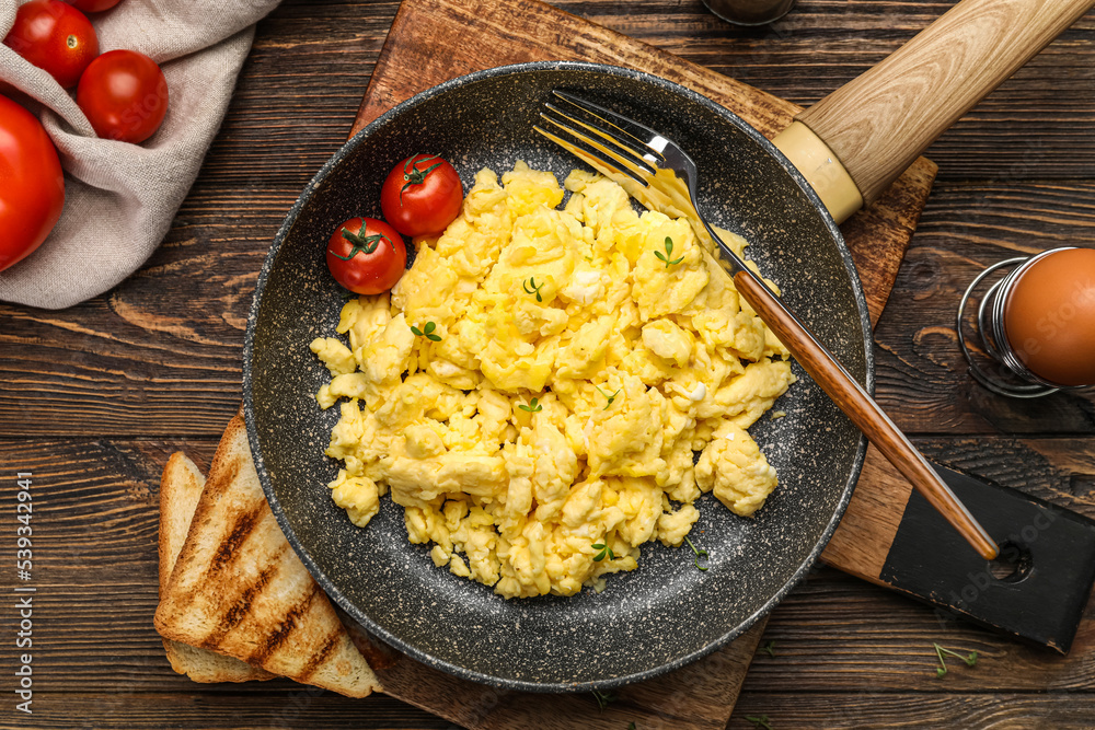 Frying pan with tasty scrambled eggs, tomatoes and toasts on wooden table