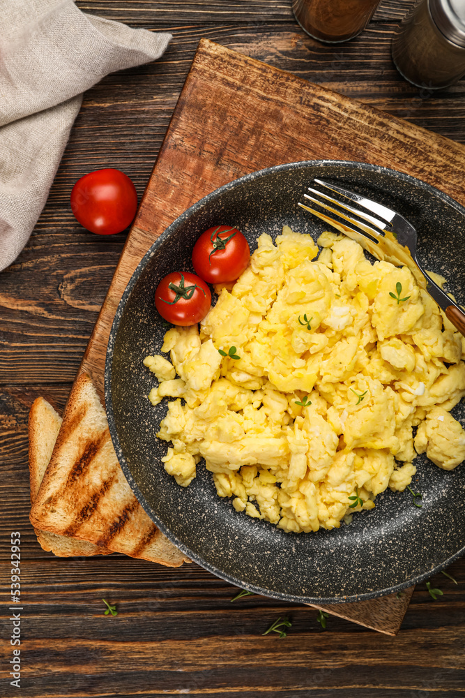 Frying pan with tasty scrambled eggs, tomatoes and toasts on wooden table, closeup