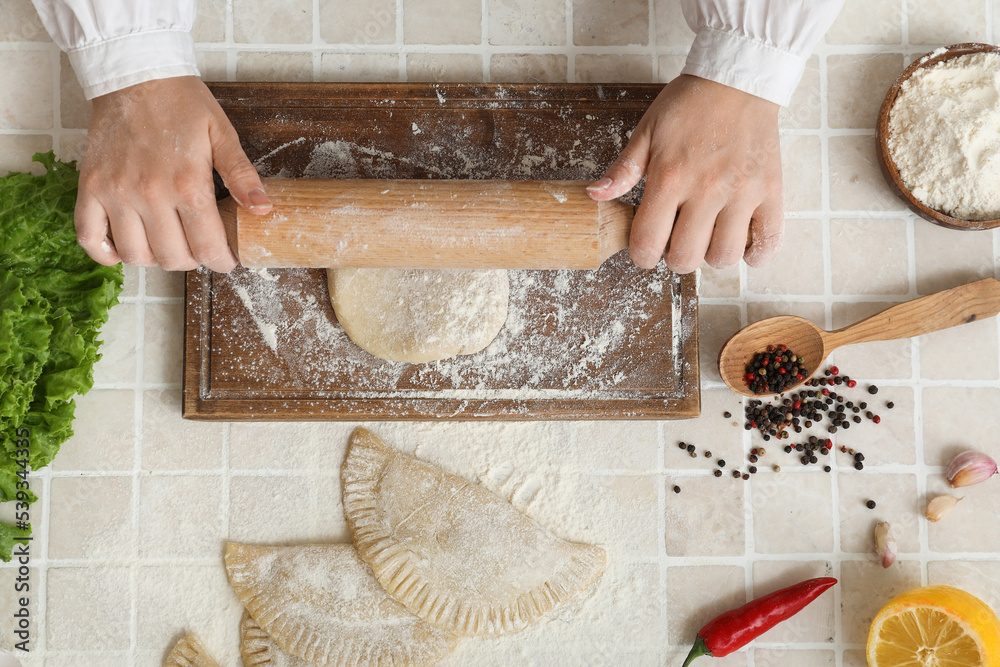 Woman rolling out dough for chebureks on beige tile background, top view