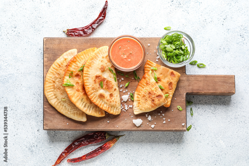 Wooden board with tasty chebureks, sauce, green onion and chili peppers on light background