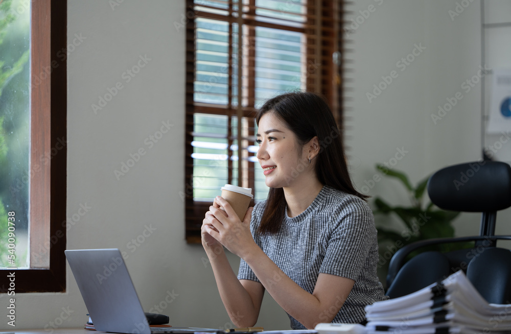 美丽的年轻亚洲女商人拿着咖啡杯和笔记本电脑在办公室工作，面带微笑。