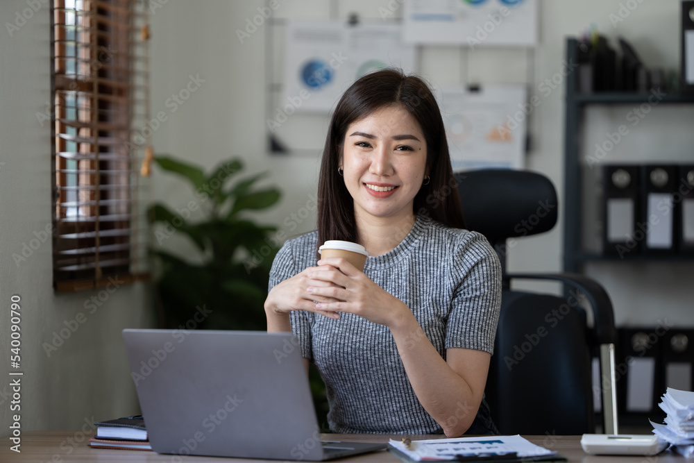 Beautiful young Asian businesswoman smiling holding a coffee mug and laptop working at the office.