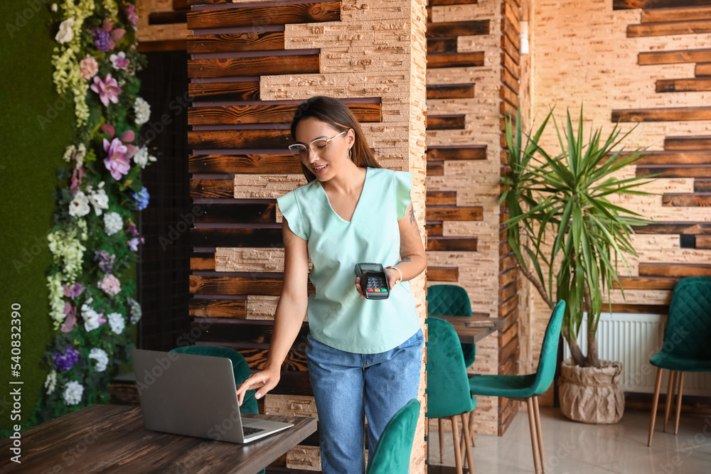 Beautiful business owner with payment terminal and laptop in her cafe