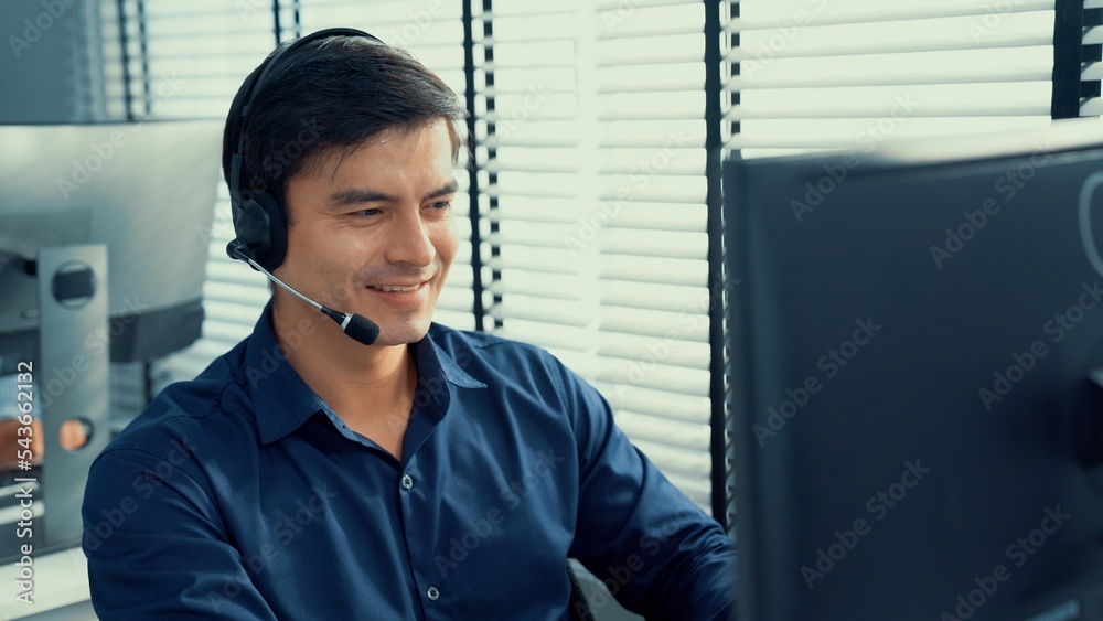 Young competent asian male call center agent working at his computer while simultaneously speaking w