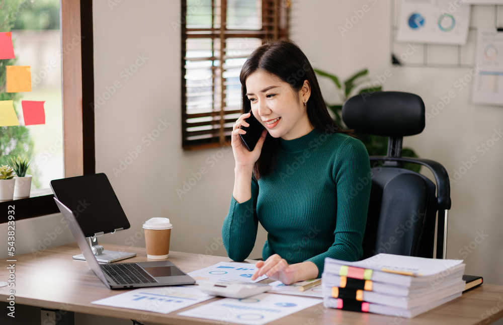 亚洲商务女性坐在办公室里微笑着打电话和使用笔记本电脑。