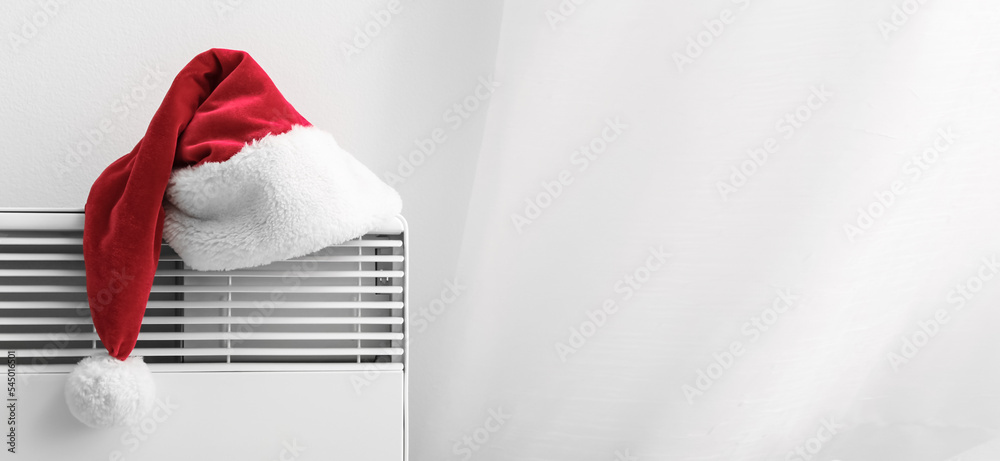 Santa hat on electric radiator near light wall with space for text