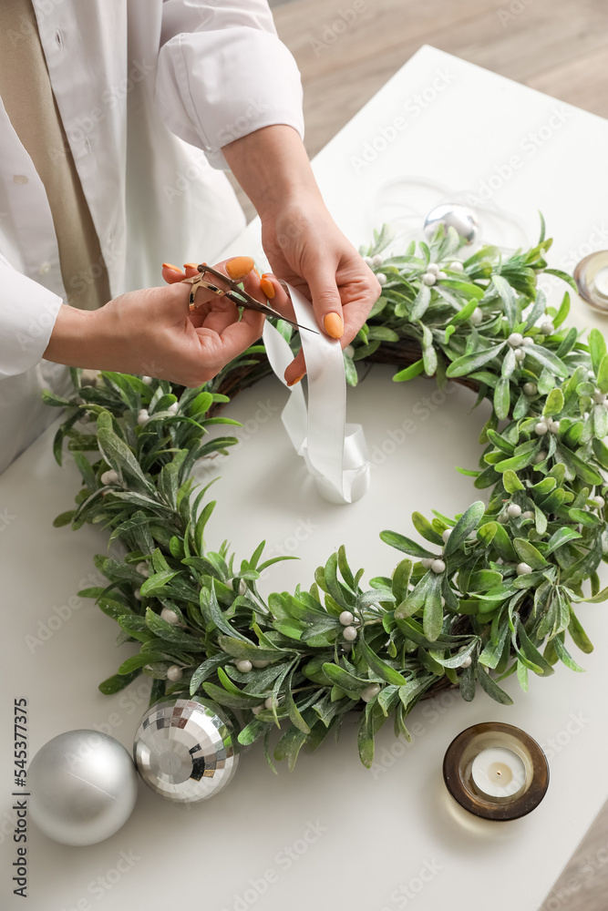 Woman cutting ribbon for Christmas mistletoe wreath at white table, closeup