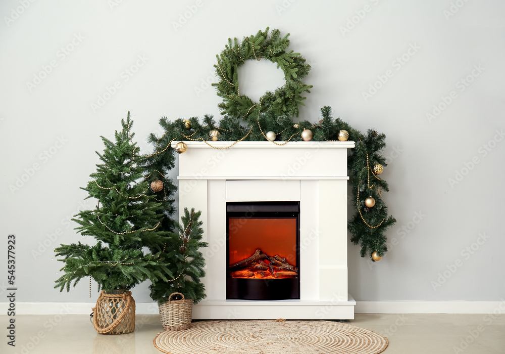 Electric fireplace with Christmas trees, wreath and fir branches near light wall