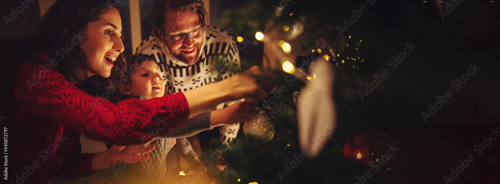 Happy family with mother, father, and child decorate Christmas tree in a cozy warm family home durin