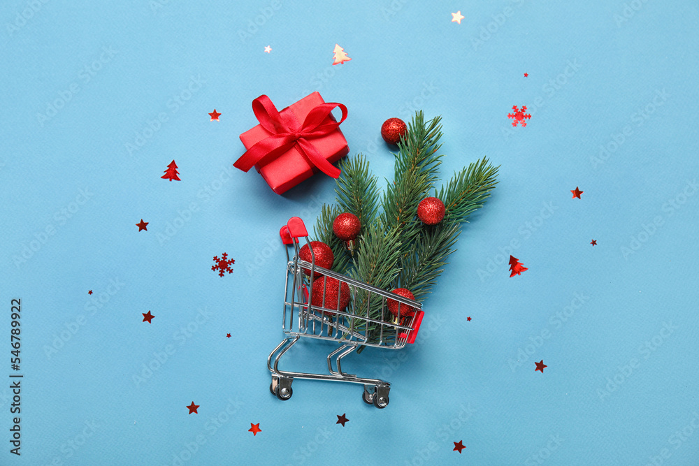 Shopping cart with fir branches, Christmas balls and gift on blue background