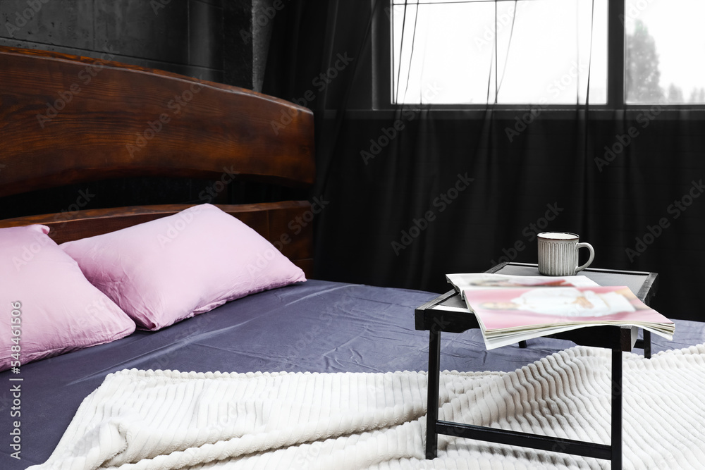 Tray with magazine and cup on comfortable bed in dark room