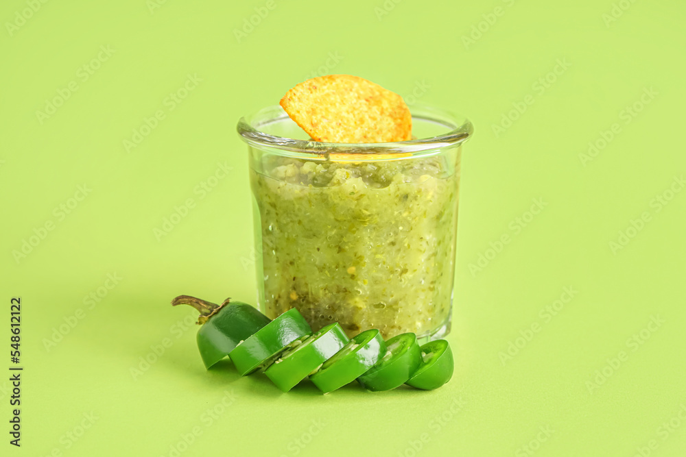 Bowl of tasty green salsa sauce and jalapeno peppers on green background