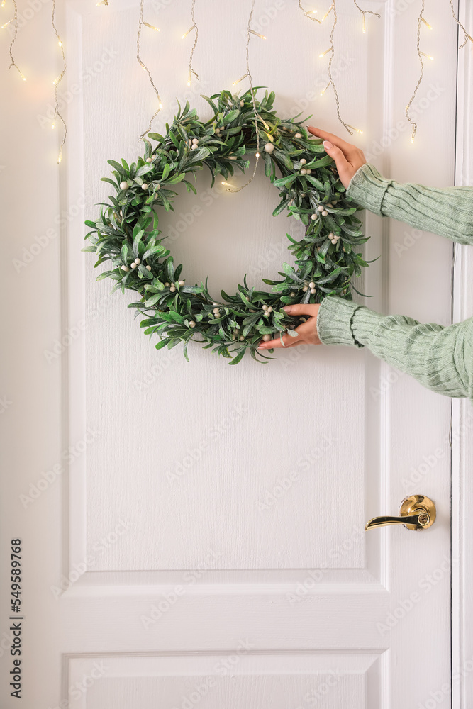 Woman hanging Christmas mistletoe wreath on white door in hall