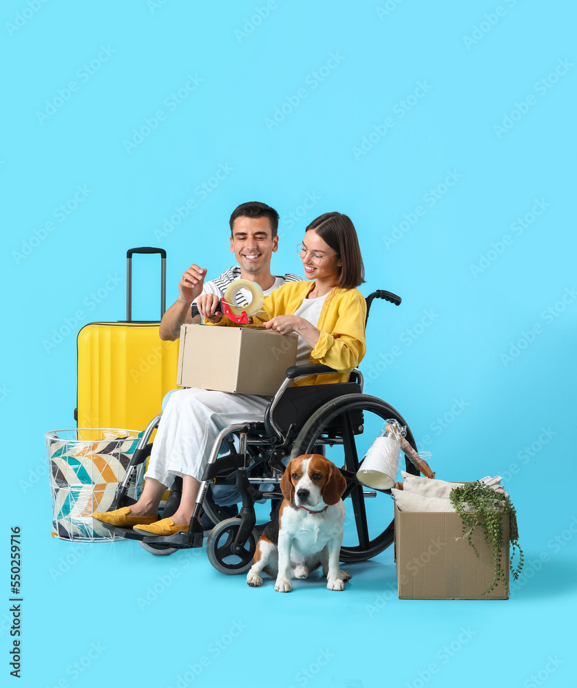 Young man with his wife in wheelchair, moving boxes and dog on blue background