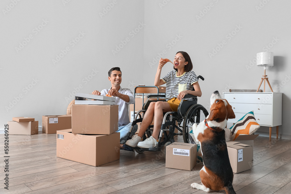 Young man with his wife in wheelchair and dog eating pizza in their new flat on moving day