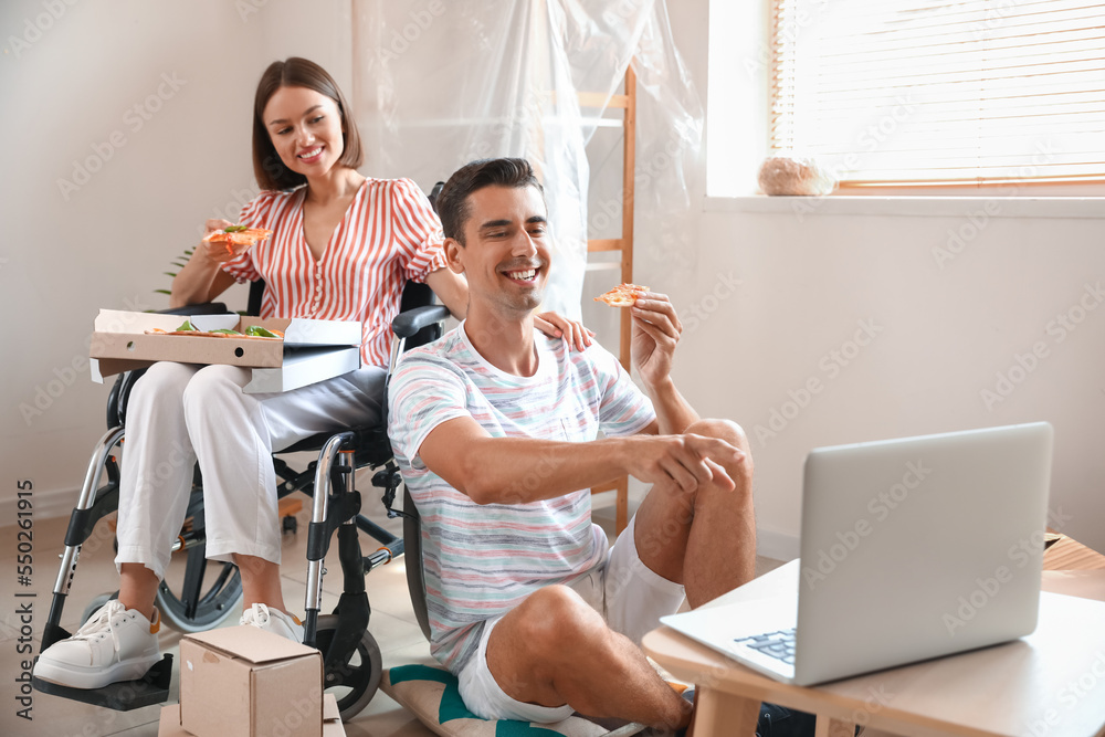 Young man and his wife in wheelchair with pizza watching video in room on moving day