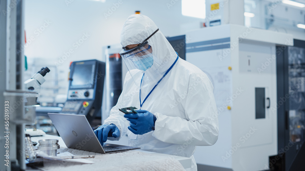 Modern Technology Concept: Engineer in Coverall Working on Laptop Computer, Examining a Circuit Boar