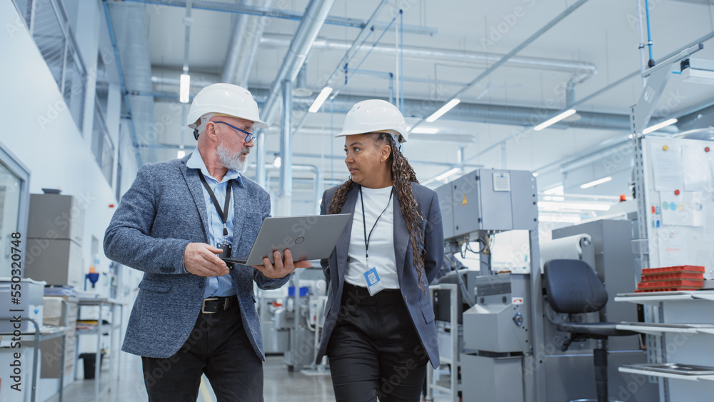 Portrait of a Two Diverse Heavy Industry Engineers in Hard Hats Walking with Laptop Computer and Tal