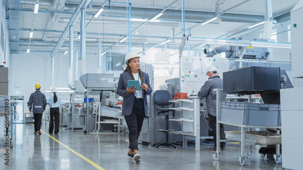 Portrait of a Black Female Engineer in Hard Hat Walking and Using Laptop Computer at Electronic Manu