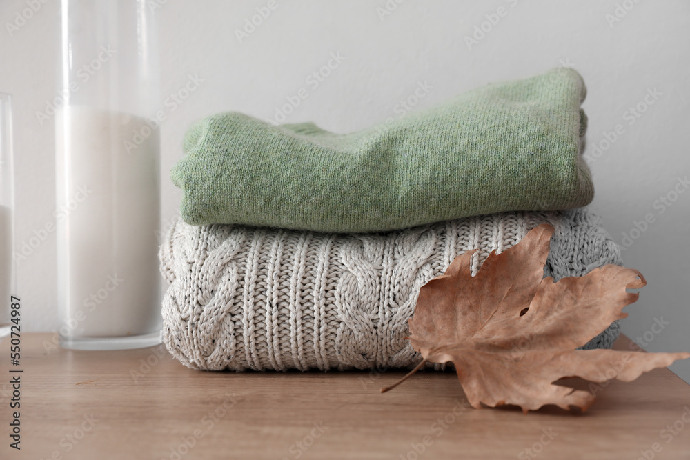 Folded sweaters with fallen leaf on table near light wall, closeup