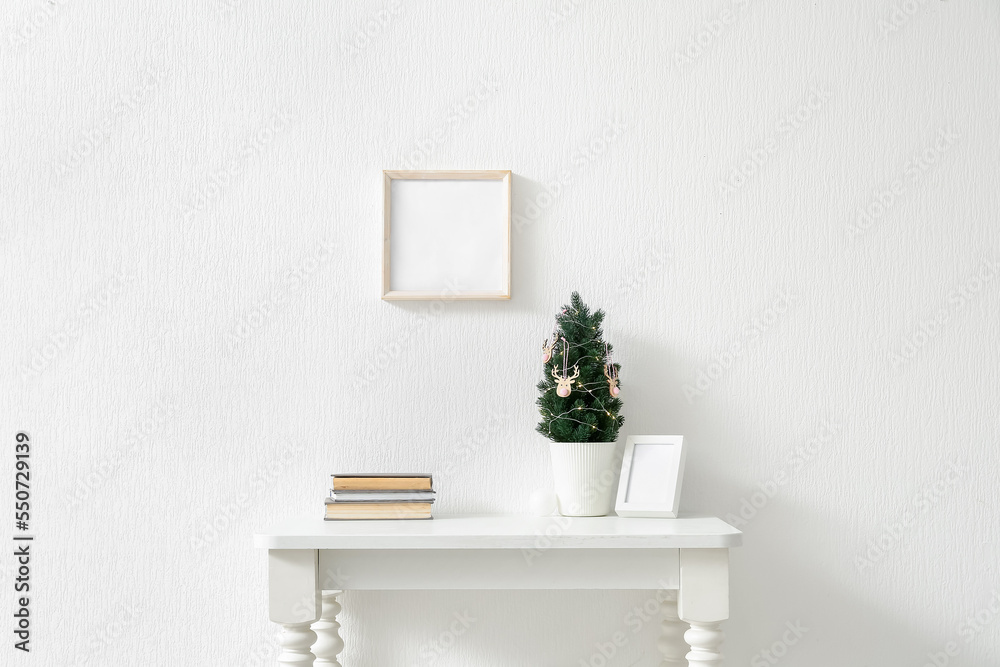 Small Christmas tree, frame and books on table near light wall