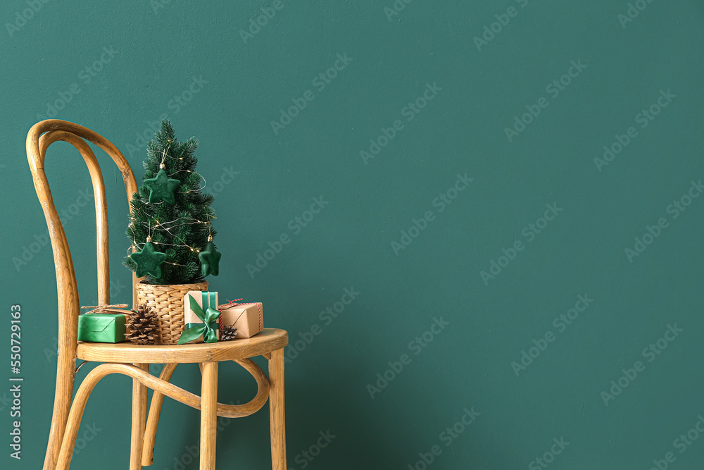 Small Christmas tree with pine cones and gifts on chair near green wall