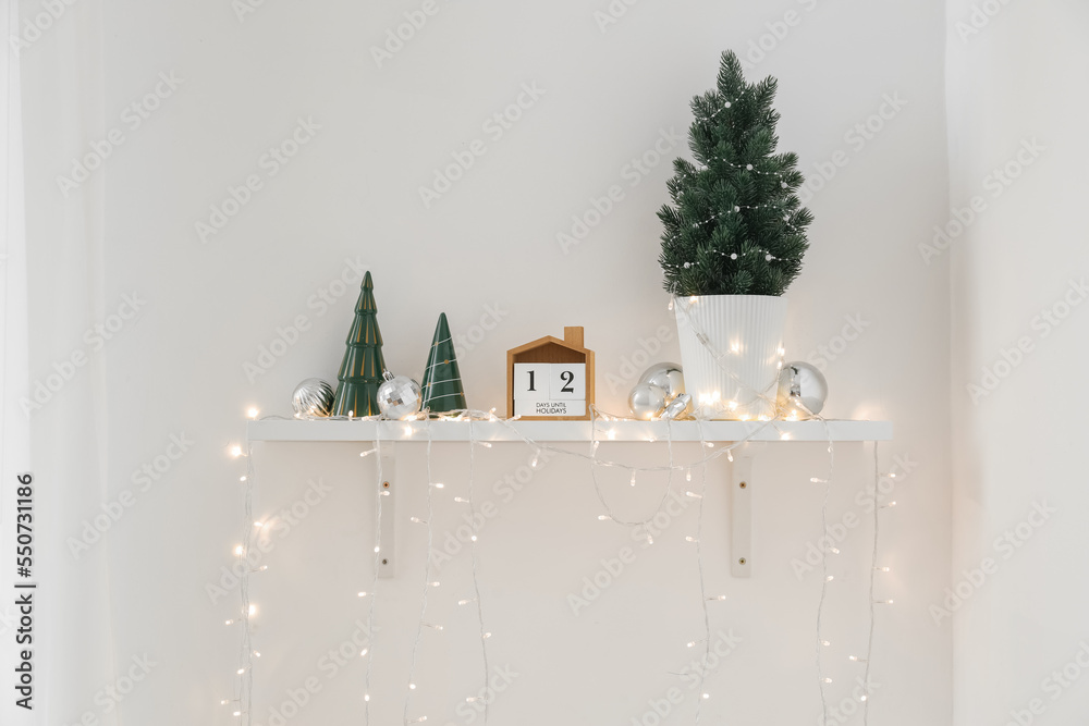 Shelf with small Christmas tree, calendar, toys and glowing lights on light wall