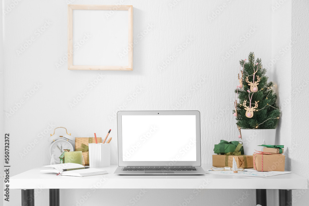 Laptop with Christmas tree and presents on table near light wall