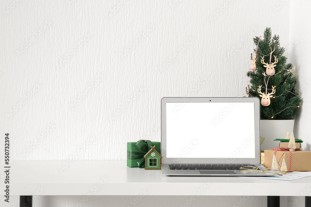 Laptop with Christmas tree and presents on table near light wall