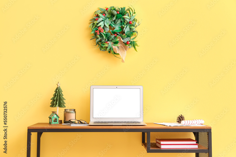 Laptop with Christmas toys and candle on table near yellow wall