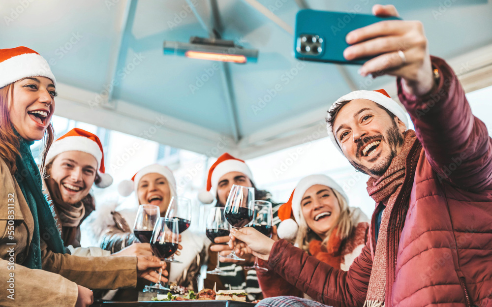 Young friends wearing santa claus hats taking selfie with smartphone sitting at dining restaurant ta