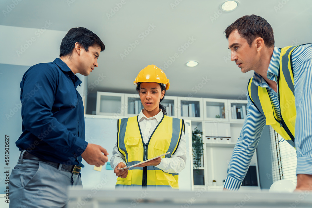 A team of competent engineers wearing safety equipment is working on blueprints with a tablet while 