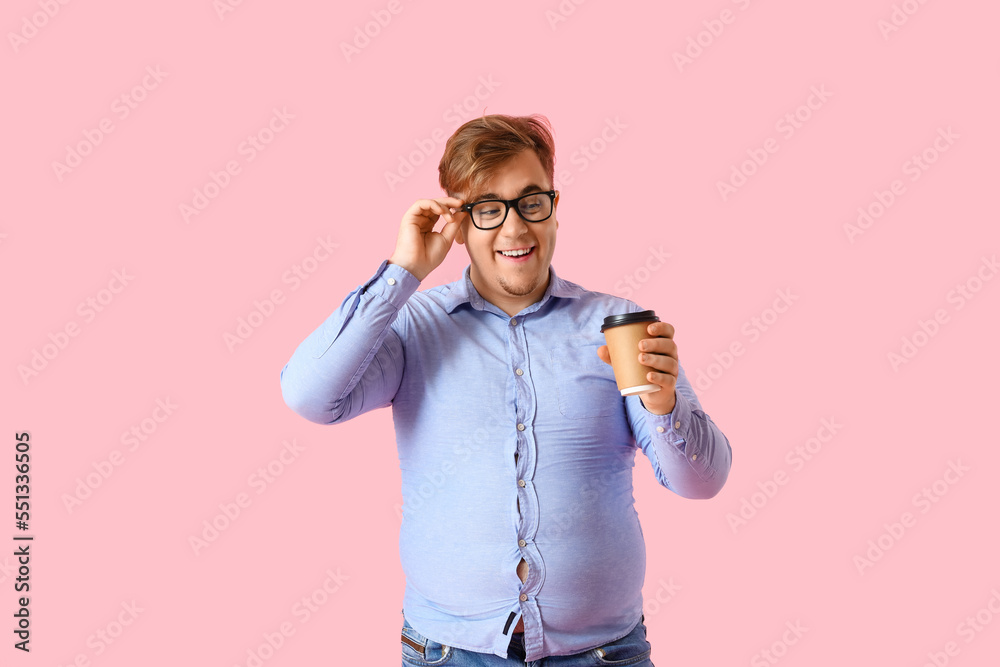 Young overweight man in tight shirt with cup of coffee on pink background